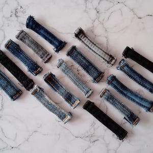 Może przedstawiać: Asortyment pasków z tkaniny dżinsowej w różnych odcieniach niebieskiego i czarnego, ułożonych na marmurowej powierzchni. Paski, wycięte z dżinsów, mają widoczne szwy i postrzępione krawędzie. Układ jest ukośny, tworząc wzór. Paski mają około 10 cm długości.