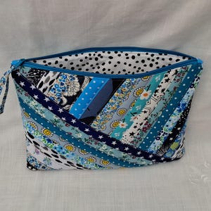 May include: A zipped pouch featuring a patchwork design of diagonal fabric strips in various shades of blue, white, and black. The exterior displays patterns including stars, flowers, and smiley faces. The interior lining is white with black dots.