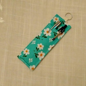 May include: A rectangular fabric pen case in teal featuring a white daisy pattern with yellow centers. The case includes a silver metal key ring at the top and contains two pens, one silver and one black, inserted into the open top pocket.
