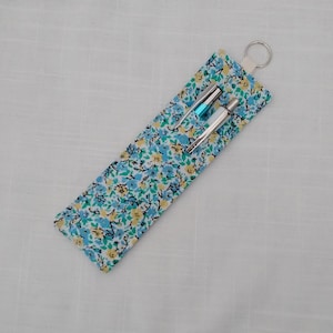 Op de afbeelding: Een stoffen etui met bloemenpatroon in blauw, geel en wit op een turquoise achtergrond. Het bevat twee zilveren en turquoise pennen en heeft een sleutelhanger. De etui is ongeveer 20 cm lang.