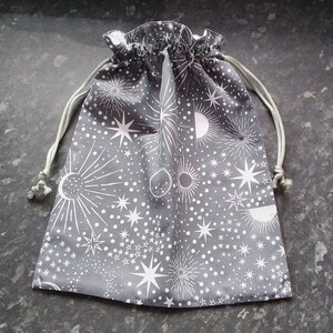 May include: A grey drawstring bag with a white star and moon pattern. The bag is made of fabric and has a white drawstring.