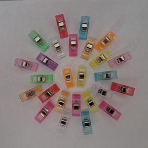 Peut inclure: Un ensemble de pinces de couture colorées dans des teintes translucides variées, notamment rose, vert, orange et bleu. Chaque pince est dotée d'un fermoir en métal argenté. Les pinces sont disposées en cercle.