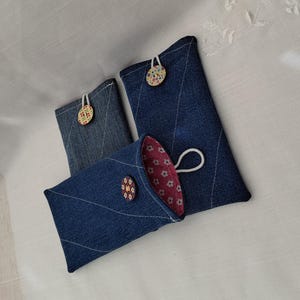 May include: Three denim pouches with floral-patterned buttons and white string closures. The pouches are various sizes, with a quilted diamond pattern. One pouch is open, revealing a pink floral interior.