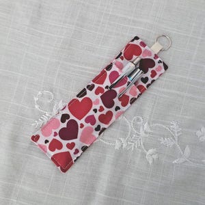 Peut inclure: Un étui à stylos en tissu orné de cœurs rouges, roses et bordeaux. Il contient deux stylos et possède une petite boucle avec un anneau métallique. Le fond est un tissu blanc texturé.