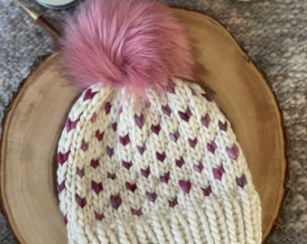 May include: White knitted hat with a pink pom pom and a brown leather tag that says "Knit with Heart". The hat has a pattern of pink hearts.