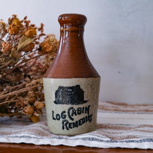 May include: A ceramic bottle with a narrow neck and a wide base. The top half is a glossy brown, while the bottom half is a speckled cream color. A black graphic of a log cabin is printed above the text "LOG CABIN REMEDIES" in black lettering.