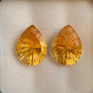 May include: Two faceted, teardrop-shaped gemstones with a warm, golden-yellow hue. The gems are displayed in a black box with a white interior. The facets create a starburst pattern, enhancing the stones' brilliance. Ideal for jewelry making.