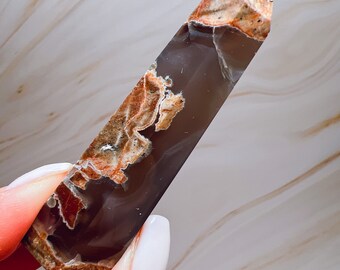 May include: A polished, elongated crystal point with a flat, angled top. The crystal features a mix of dark brown, tan, and cream colours with intricate patterns. The crystal is held by a person with white nail polish.