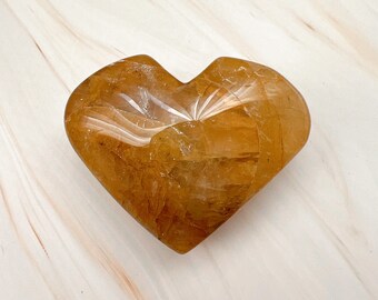 May include: A heart-shaped, amber-colored crystal. The stone has a translucent quality with visible internal patterns and a polished surface. The heart is set against a white and beige marbled background. This decorative piece is ideal for home decor or as a unique gift.