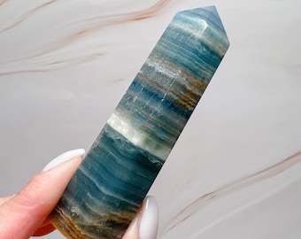 May include: A blue and white banded crystal tower with a pointed top. The crystal has layers of blue, white, and brown hues. The crystal is held by a person with a light-colored background.