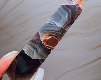 May include: A banded agate crystal point in shades of grey, brown, and black. The crystal is polished and has a pointed top. The background is a marble-effect surface.