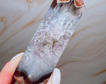 May include: A rectangular crystal tower with a polished finish. The top section is reddish-brown, leading down to bands of grey, white, and lavender. The crystal is held against a marbled background.