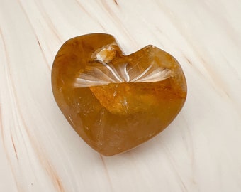 May include: A heart-shaped, amber-colored crystal sculpture with carved white lines radiating from the center. The crystal has a smooth, polished surface and sits on a light-colored, marbled background.