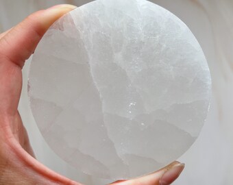 May include: A round, white selenite crystal plate with a textured surface. The plate is held in a hand, showcasing its smooth, polished appearance. This decorative piece is ideal for displaying crystals or as a unique home decor accent.