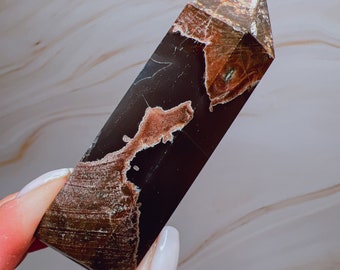 May include: A polished crystal obelisk with a dark brown base and lighter brown and tan patterns. The crystal is held by a hand with white nail polish. The obelisk has a pointed top and is a decorative item.