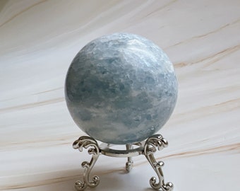May include: A light blue sphere with a textured surface, displayed on a decorative silver metal stand. The stand has ornate, curved legs. The sphere appears to be made of stone or crystal.