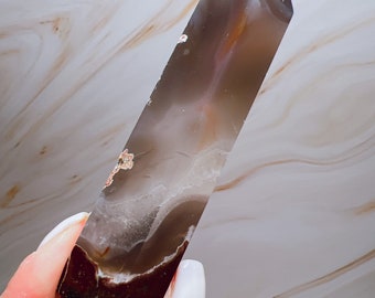 May include: A polished agate crystal obelisk. The crystal displays bands of brown, white, and grey, with a pointed top. The obelisk is held in a hand, showcasing its natural patterns and colours. This decorative crystal is a unique home decor item.