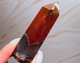 May include: A polished gemstone obelisk in reddish-brown and grey. The obelisk has a rectangular shape with a pointed top. The stone has natural patterns and colour variations, creating a unique decorative item. The obelisk is approximately 10 cm in height.