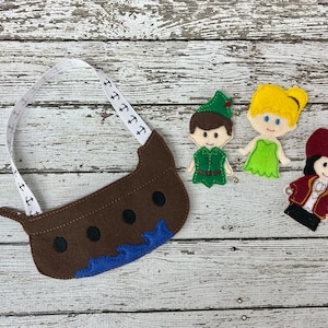 Puede incluir: Un barco de fieltro marrón con una correa de cinta blanca y anclas impresas en él. Tres títeres de dedo de fieltro: Peter Pan en verde, Campanita en verde y un pirata en rojo.