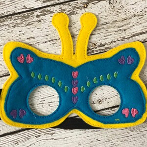 A blue felt butterfly mask with yellow edges, yellow antennae, and pink and green embroidered details lies on a white wooden surface.