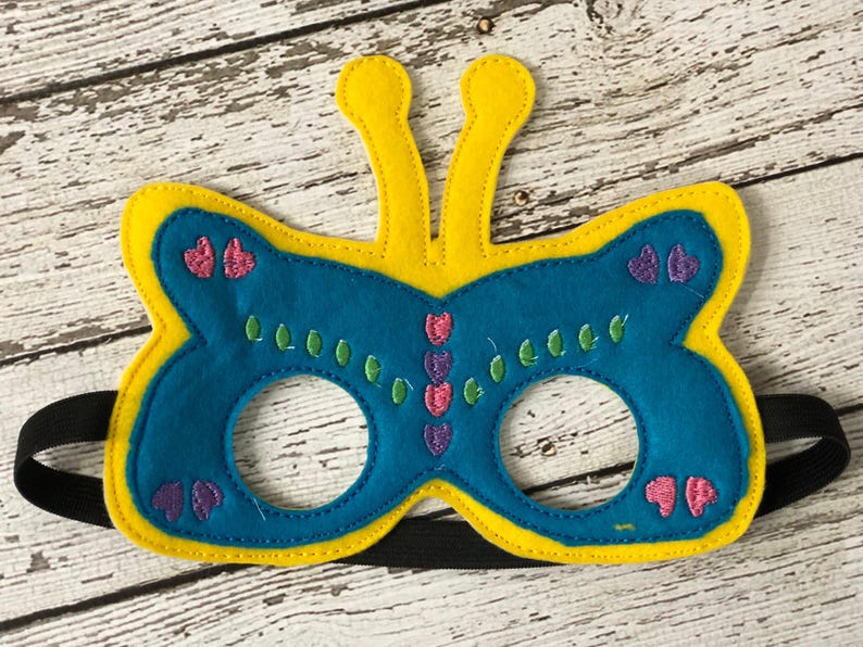A blue felt butterfly mask with yellow edges, yellow antennae, and pink and green embroidered details lies on a white wooden surface.