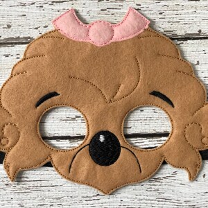 Close-up of a brown felt bear mask adorned with a small pink bow on a rustic white wood background.