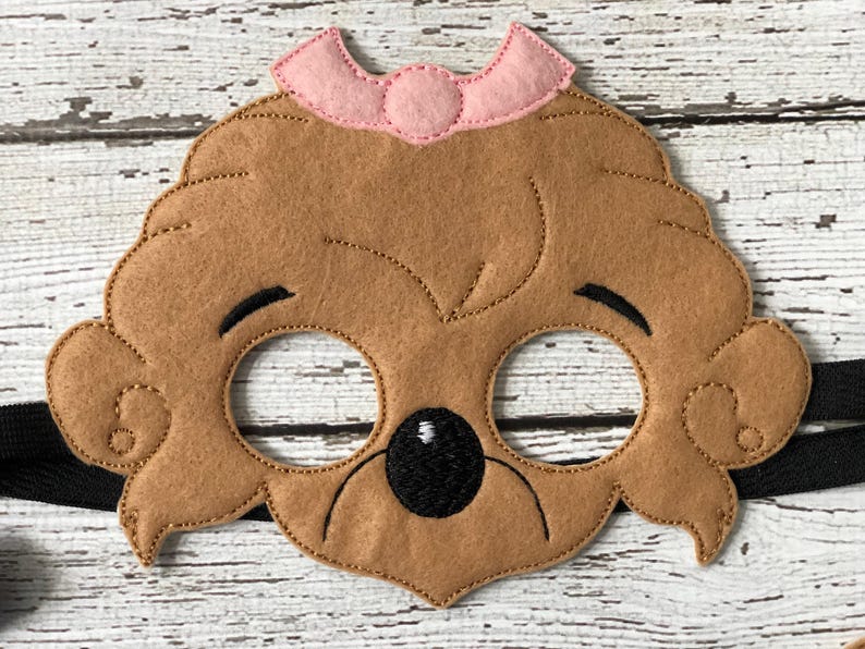 Close-up of a brown felt bear mask adorned with a small pink bow on a rustic white wood background.