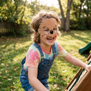 A smiling child wearing a brown bear felt mask and tie-dye shirt plays outdoors near a wooden climbing structure.