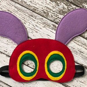 A red felt caterpillar mask with purple ears, green eye circles outlined in yellow, and a black elastic band rests on a white wood backdrop.