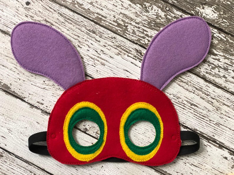 A red felt caterpillar mask with purple ears, green eye circles outlined in yellow, and a black elastic band rests on a white wood backdrop.