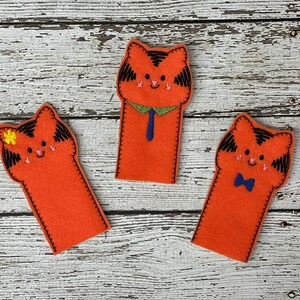 Op de afbeelding: Drie oranje vingerpoppen van vilt in de vorm van katten. Eén kat heeft een blauwe stropdas, één heeft een blauwe strik en één heeft een gele bloem.