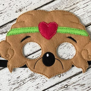 Close-up of a brown felt bear mask with a green headband and a red heart centered on a white wood backdrop.