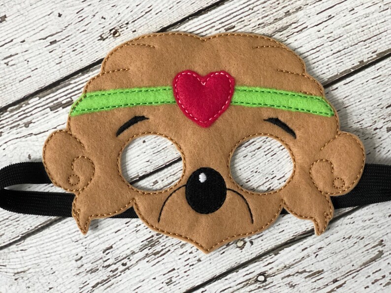 Close-up of a brown felt bear mask with a green headband and a red heart centered on a white wood backdrop.