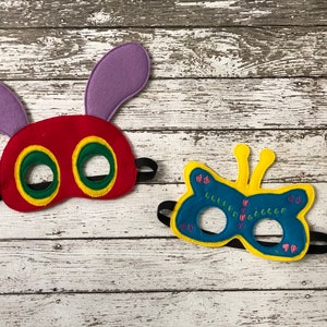 May include: Two felt masks, one red with purple antennae and green eyes, and one blue with yellow antennae and pink and green details. The masks are on a white wooden background.