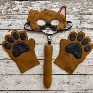 May include: A brown felt dog costume set, including a mask, two paw-shaped gloves, and a tail. The mask has a black eye covering and pink ear details. The gloves and tail feature dark gray paw prints.