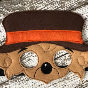 Close-up of a brown felt bear mask featuring a brown top hat with an orange band on a rustic white wood background.