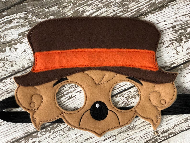 Close-up of a brown felt bear mask featuring a brown top hat with an orange band on a rustic white wood background.