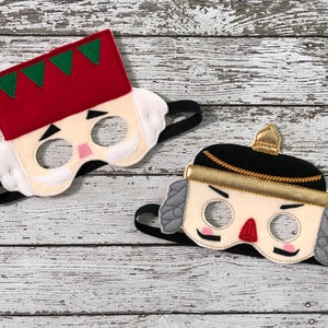 Puede incluir: Dos máscaras de cascanueces de fieltro con cintas elásticas negras. Una máscara tiene un sombrero rojo con ribetes verdes y una cara blanca. La otra máscara tiene un sombrero negro con ribetes dorados y una cara blanca.