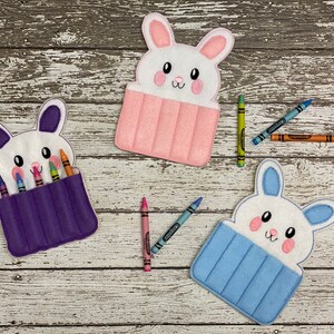 May include: Three felt bunny crayon holders. The first is purple with six crayons inside. The second is pink with no crayons inside. The third is blue with no crayons inside.