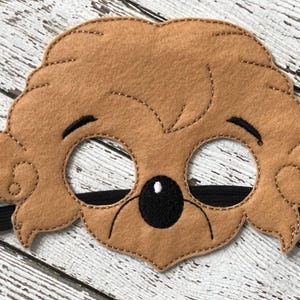 Close-up of a plain brown felt bear mask with black elastic strap on a white weathered wood surface.