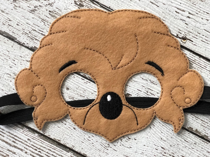 Close-up of a plain brown felt bear mask with black elastic strap on a white weathered wood surface.