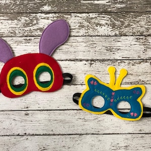 Two felt masks rest on a white wooden background, featuring a red caterpillar mask with purple ears and a blue butterfly mask with yellow trim.