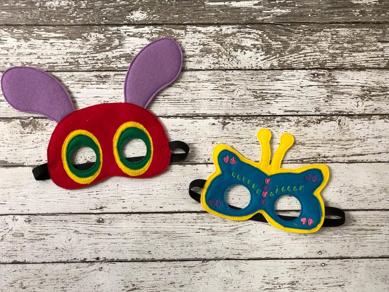 Two felt masks rest on a white wooden background, featuring a red caterpillar mask with purple ears and a blue butterfly mask with yellow trim.