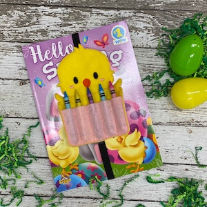 May include: A yellow felt chick with a pink pocket holding six crayons. The chick is on a book titled "Hello Spring" with a chick and egg design. The book is on a white wooden surface with green paper shreds.