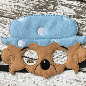 Close-up of a brown felt bear mask wearing a blue nightcap with white polka dots on a white wooden surface.