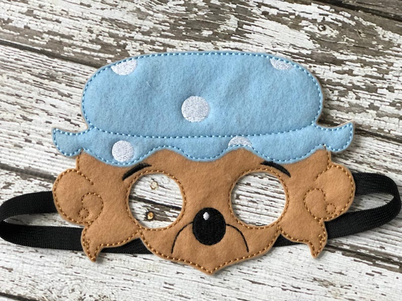 Close-up of a brown felt bear mask wearing a blue nightcap with white polka dots on a white wooden surface.