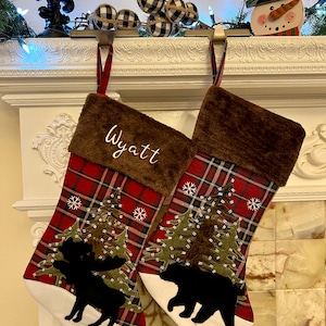 Peut inclure: Deux bas de Noël avec un motif à carreaux rouges, des hauts en fausse fourrure marron et des bas blancs. Un bas porte le nom "Wyatt" brodé. Chaque bas présente une silhouette noire d'un orignal et d'un ours avec une scène de forêt.