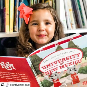 May include: A young girl with a coral bow in her hair smiles while reading a book titled "Lobo Louie and Lobo Lucy Visit the University of New Mexico." The book cover features cartoon wolves and the university's name.