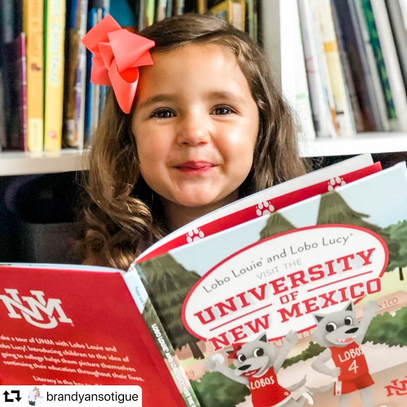 May include: A young girl with a coral bow in her hair smiles while reading a book titled "Lobo Louie and Lobo Lucy Visit the University of New Mexico." The book cover features cartoon wolves and the university's name.