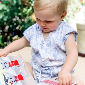 May include: A young child wearing a blue and white floral romper is engrossed in a colorful book. The book features illustrations of cartoon wolves in red jerseys with the word "LOBOS". A blue bow adorns the child's hair.